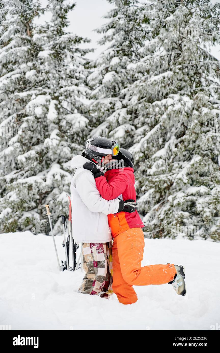 Junges Paar von Touristen küssen im Schnee gegen hypnotisierende verschneite Fichtenwald auf dem Hintergrund. Lebendige bunte Anzüge auf weißem Schnee. Konzept des Skifahrens und Beziehungen. Stockfoto