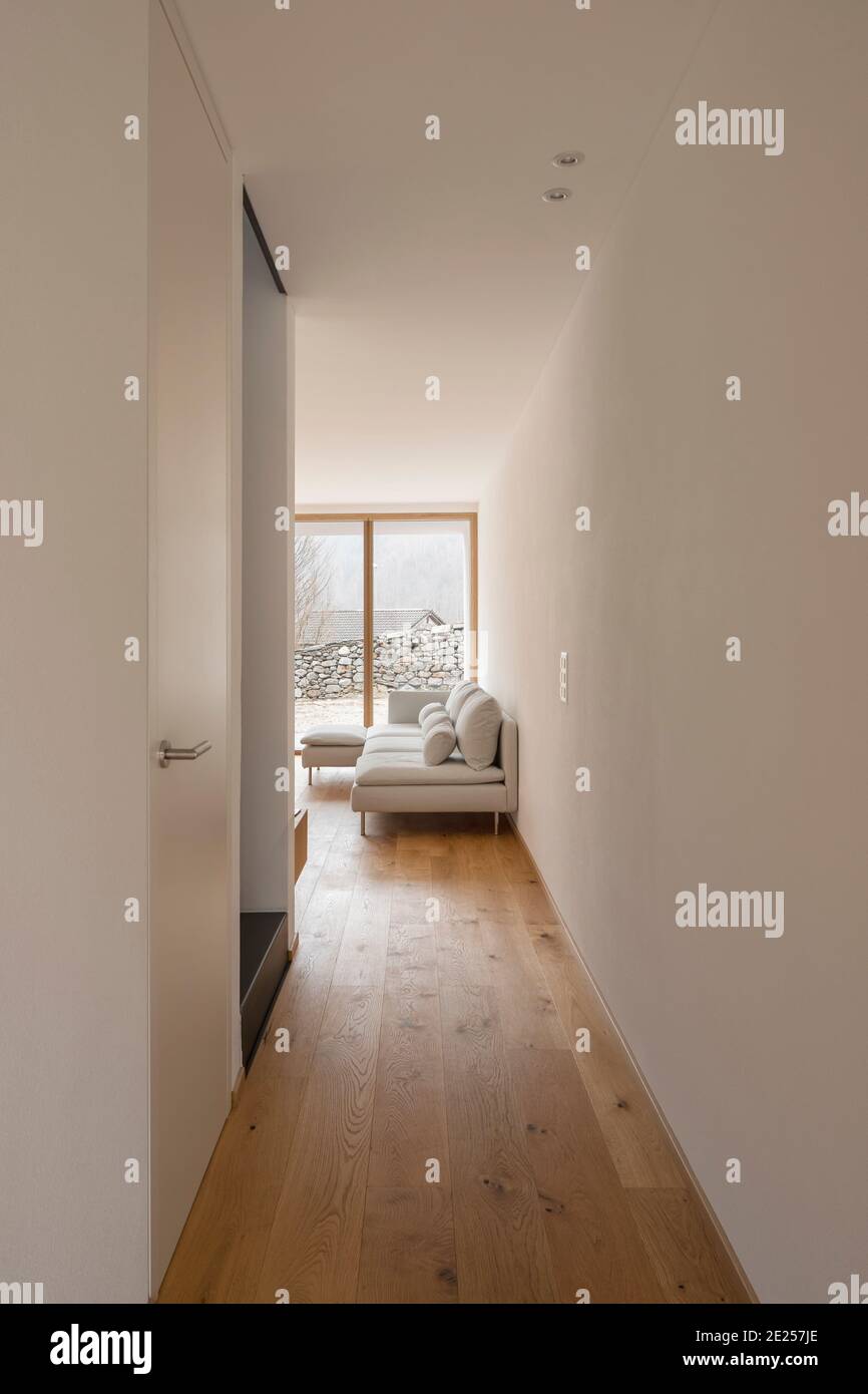 Moderne Inneneinrichtung mit weißen Wänden und Hartholzböden. Flur mit Treppe. Wohnzimmer mit Sofa und großem Fenster zur Natur. Niemand drinnen Stockfoto