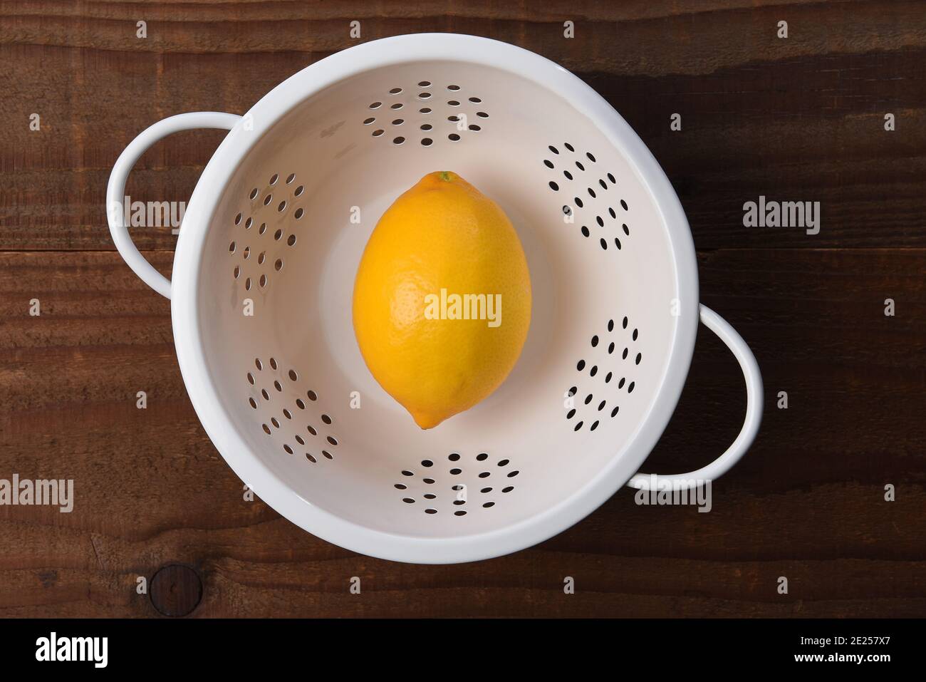 Eine einzige Zitrone in einem weißen Sieb auf einem Küchentisch aus dunklem Holz. Aufgenommen von direkt oben. Stockfoto