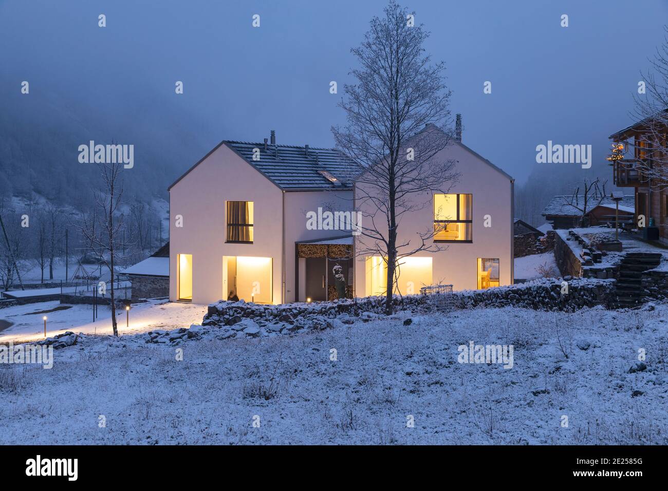 Modernes weißes Haus mit hellen Fenstern umgeben von Bergen, Schnee und Bäumen. Chirstmas Stimmung. Niemand drinnen. Stockfoto