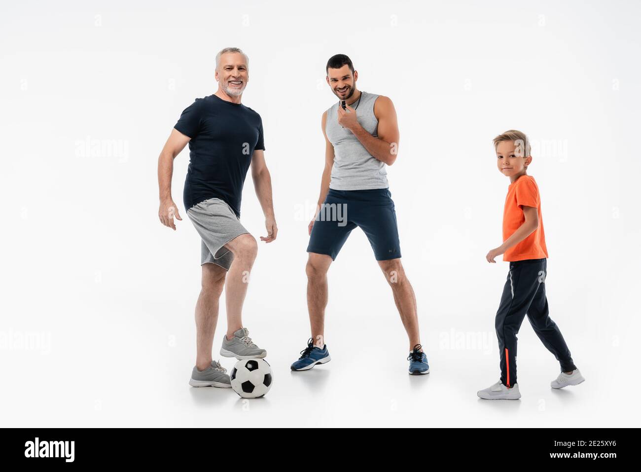 Fröhlicher Mann pfeift neben Junge und Großvater tritt auf Fußball Ball auf Weiß Stockfoto