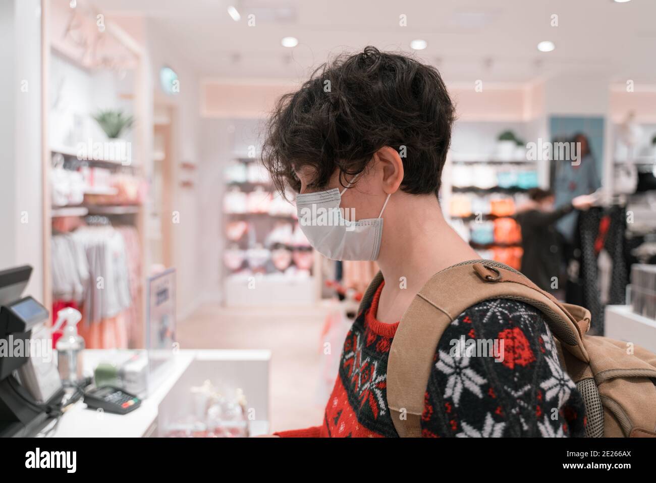 Shopping Frau trägt medizinische Maske kurze Haare. Warten an der Kasse zu zahlen. Für weihnachten oder Neujahr. Hässliche weihnachtsjacke und Rucksack. coro Stockfoto