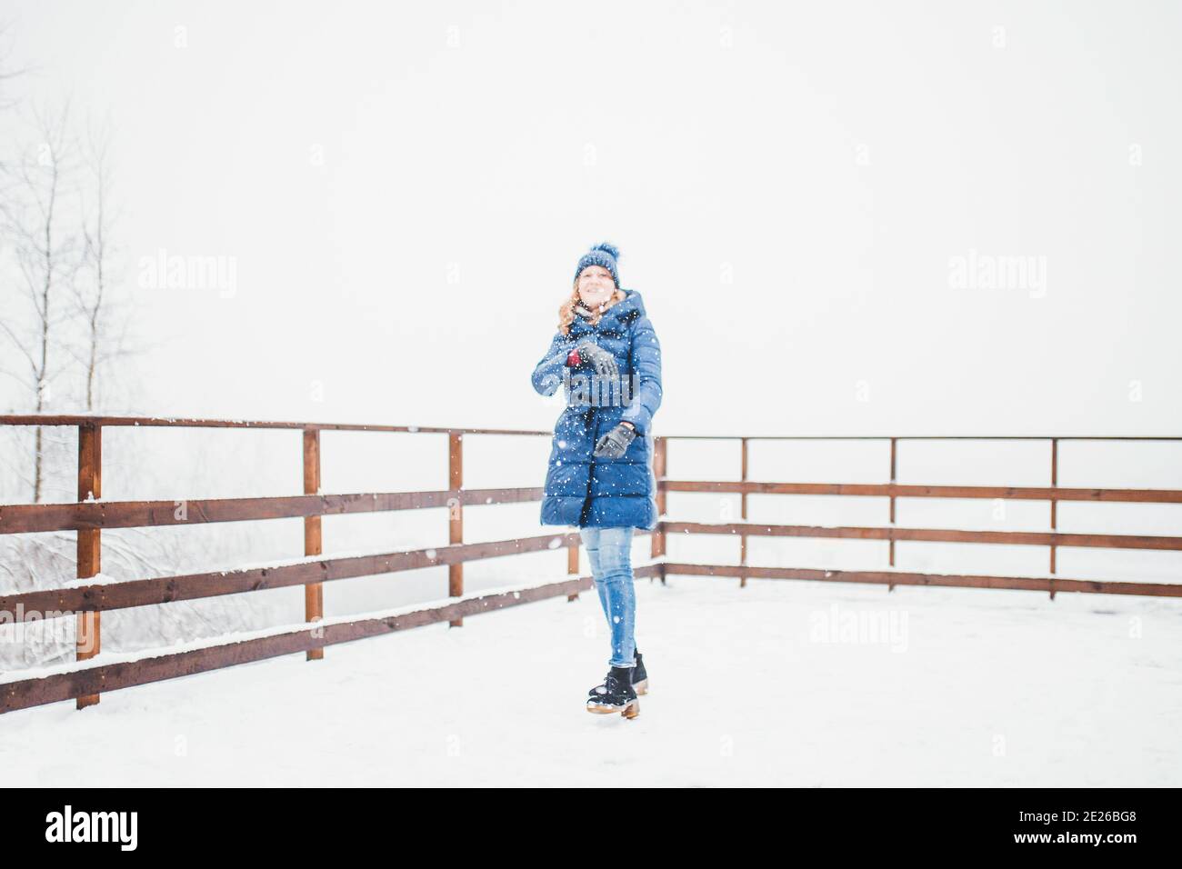 Eine hohe schlanke Blondine in einer langen blauen Daunenjacke Und eine Strickmütze im Winter auf weißem Schnee wirft Schneebälle an der Kamera - Schneeflocken fliegen - BL Stockfoto