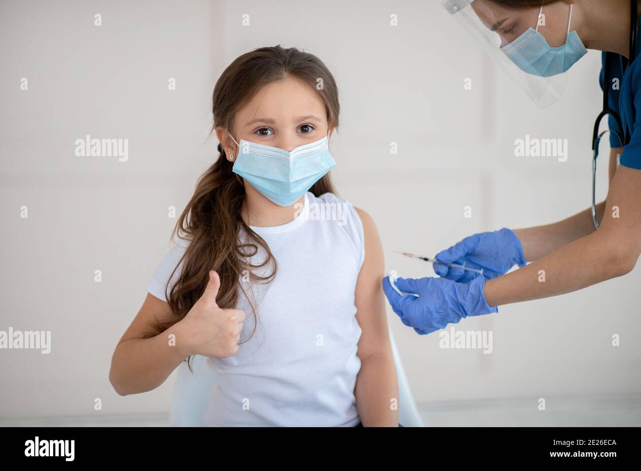 Kleines Mädchen im Gesicht Maske gestikulierenden Daumen nach oben beim Empfangen Covid-19 Impfstoffinjektion im Gesundheitszentrum Stockfoto