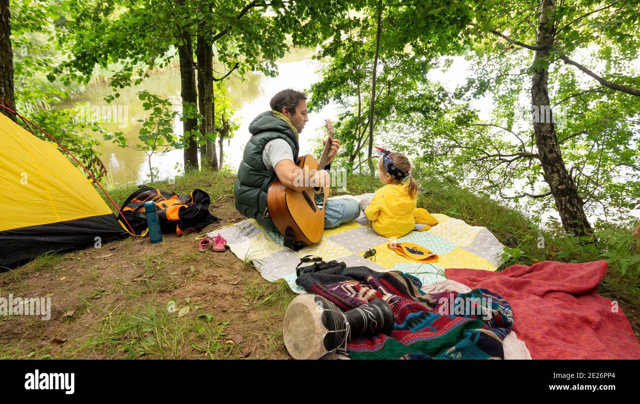 Ein Vater spielt für seine kleine Tochter Gitarre, während er auf dem Familiencamping am Ufer eines Sees Urlaub macht. Ruhe während der touristischen Trekking um Stockfoto