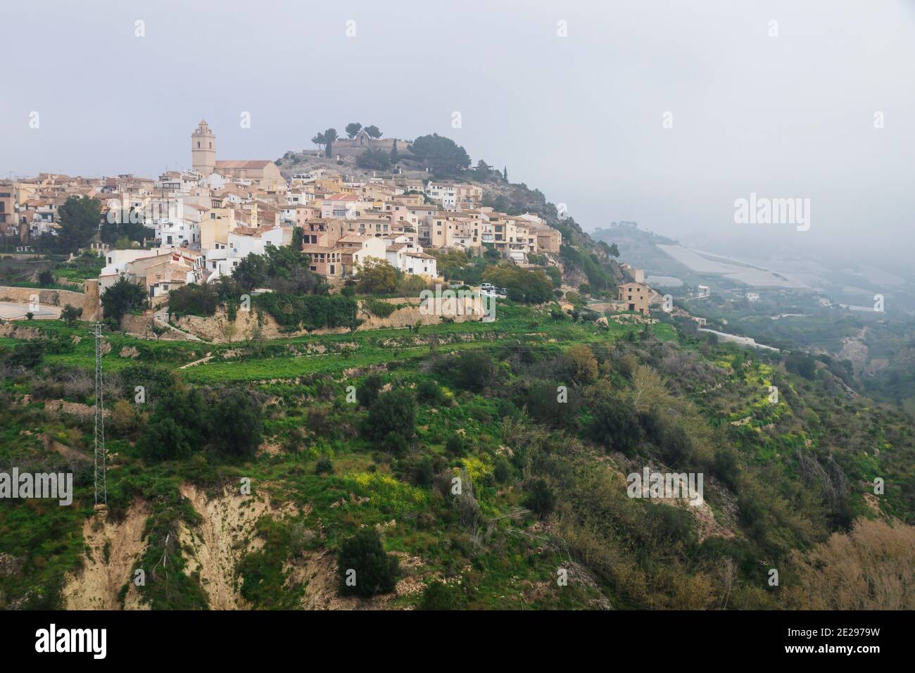 Nebliger Morgen an den Hügeln von Polop de Marina mit Kirche und castell über grünem Wald an der Costa Blanca, Spanien Stockfoto