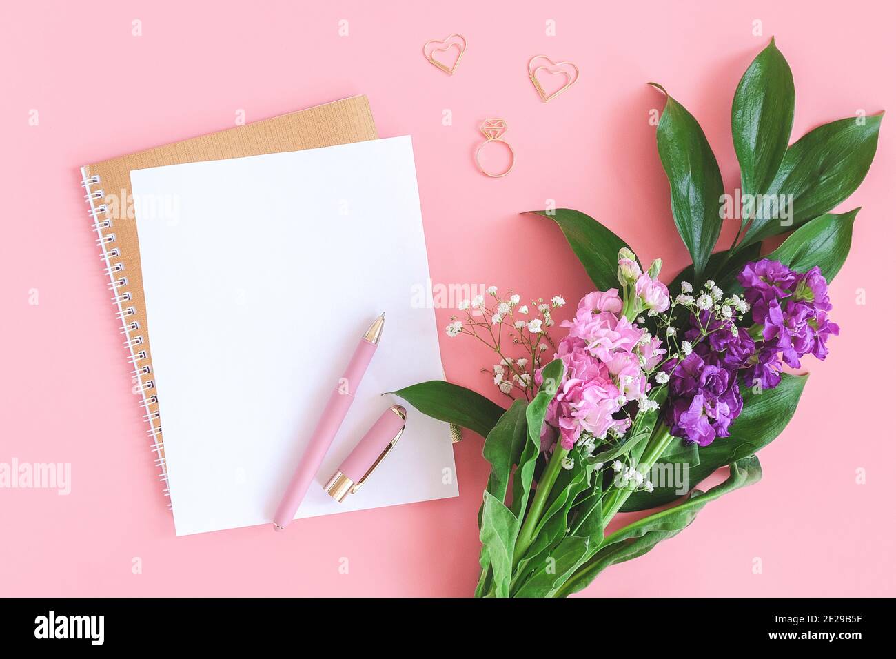 Weißes Blatt auf spiralförmigem goldenem Notizblock mit Büroklammer Herz und Ring und Blumenstrauß auf rosa Hintergrund. Konzept romantische Liebe Mockup für Stockfoto