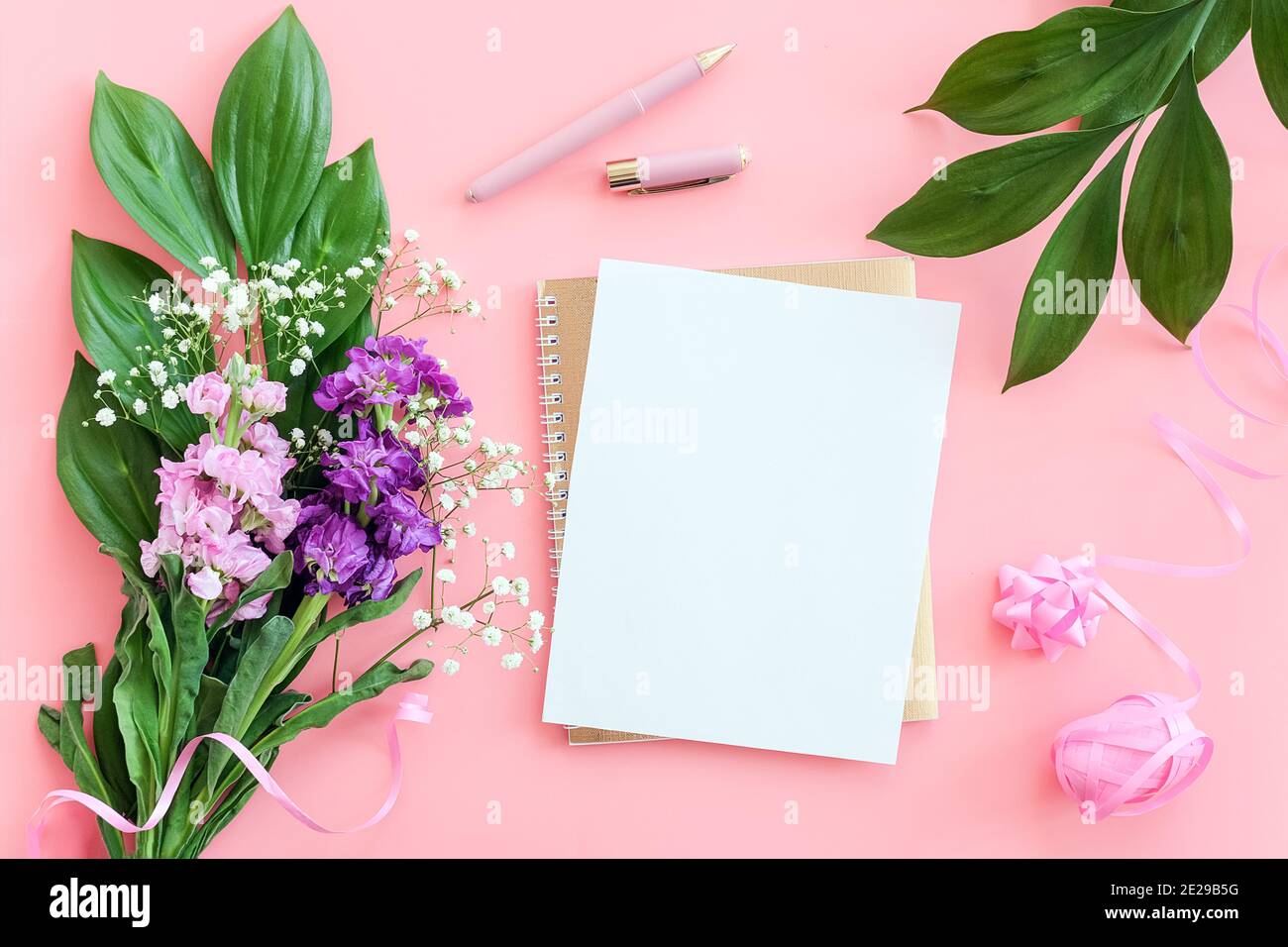 Blankes weißes Blatt auf spiralförmigem goldenem Notizblock mit Stift für Ihren Text oder Ihr Design und Blumenstrauß auf rosa Hintergrund. Konzept weiblicher Arbeitsbereich Mockup Stockfoto