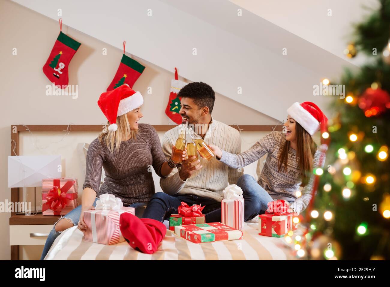 Glückliche Freunde tauschen Weihnachtsgeschenke aus, während sie im Schlafzimmer auf dem Bett sitzen. Weihnachtsferien Konzept. Stockfoto