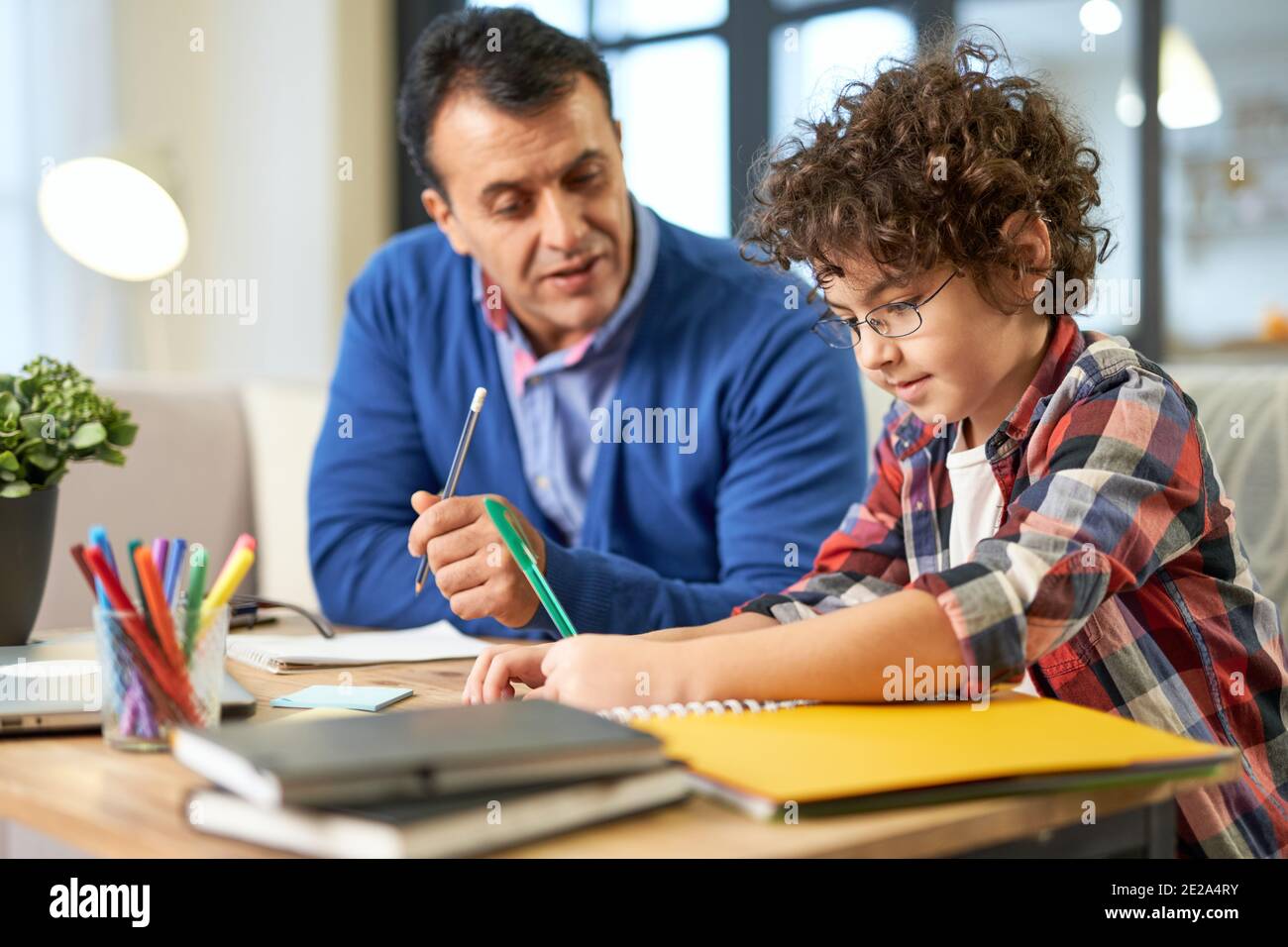 Fürsorglicher lateinischer Vater mittleren Alters, der hilft, Hausaufgaben mit seinem Sohn, Schulkind besprechen, während er zusammen am Schreibtisch zu Hause sitzt. Online studieren, Familie, Vaterschaft Konzept Stockfoto