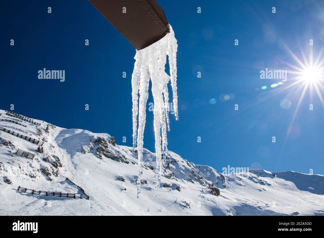 Eisstalaktiten hängen an einer Rinne in den Bergen, am blauen Himmel scheint die Sonne mit ihren Strahlen. Konzept über Eiszeit und Klimazeiten. Schnee. Stockfoto