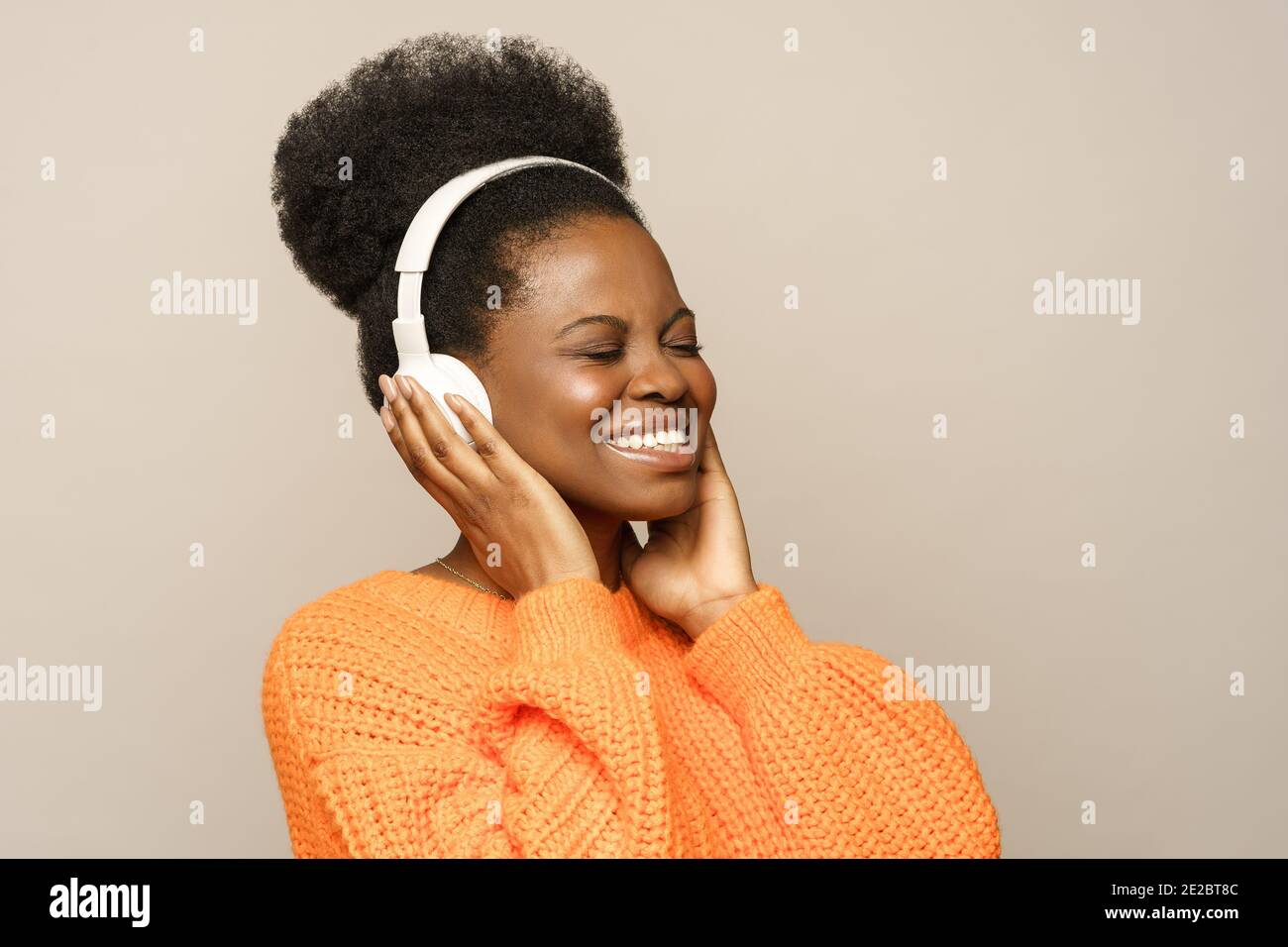 Lächelnde Afro-Frau in orange Jumper berühren drahtlose Kopfhörer, hört Lieblingsmusik isoliert Stockfoto