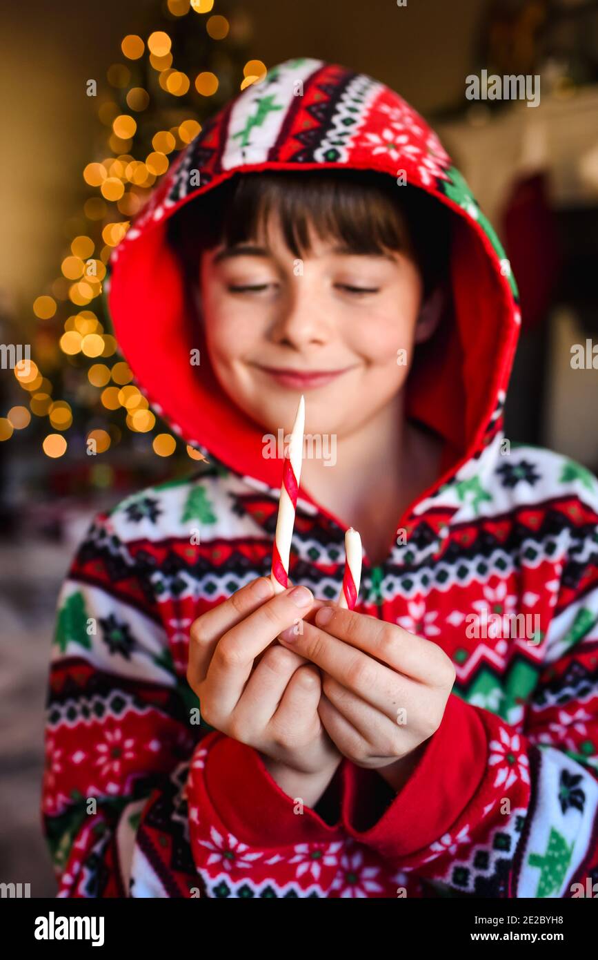 Süßer Junge im festlichen Pyjama, der zu weihnachten einen Zuckerrohr isst. Stockfoto