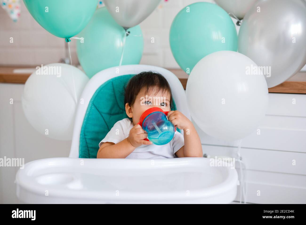 Kleiner Junge, der zu Hause in einem blauen Hochstuhl sitzt Auf der weißen Küche und spielen mit Holzlöffel auf Hintergrund mit Ballons Stockfoto