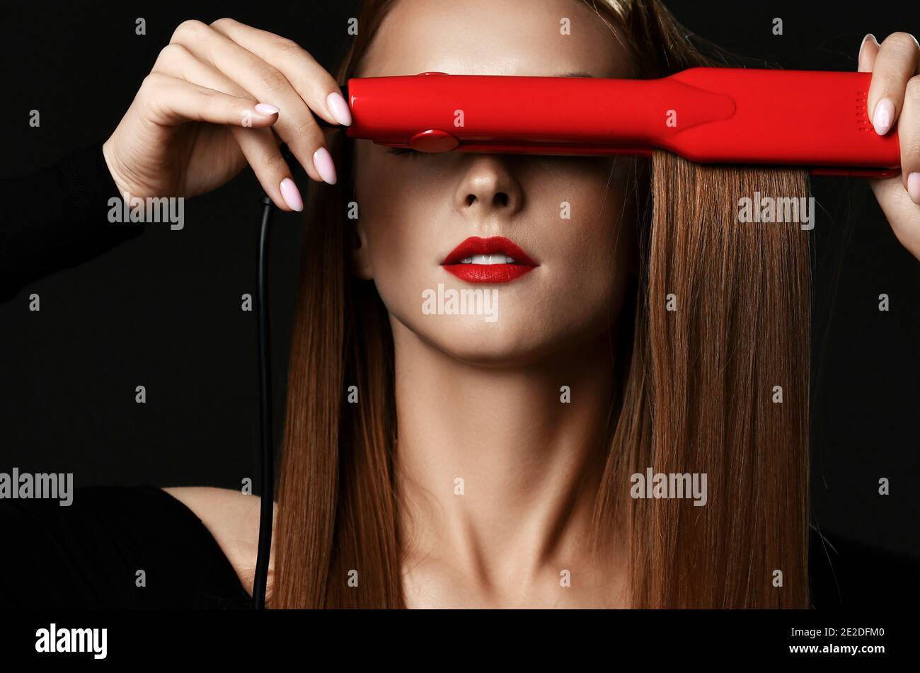 Junge hübsche Frau mit langen seidig geraden Haaren in schwarz Kleidung, die die Augen mit rotem Haarglätter bedeckt Stockfoto