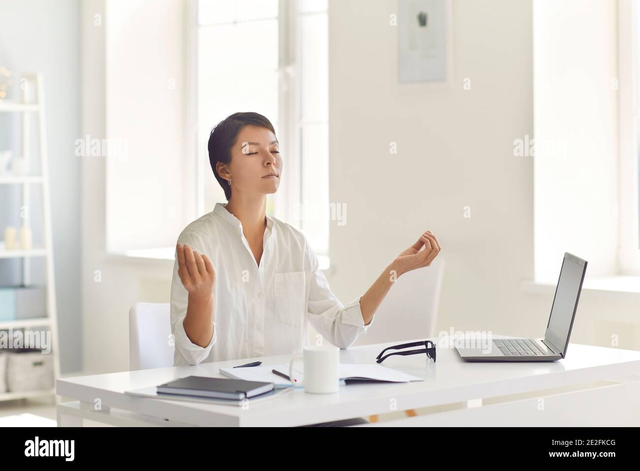 Geschäftsfrau, die am Schreibtisch in der Nähe des Laptops sitzt und während der Meditation meditiert Arbeitstag Stockfoto