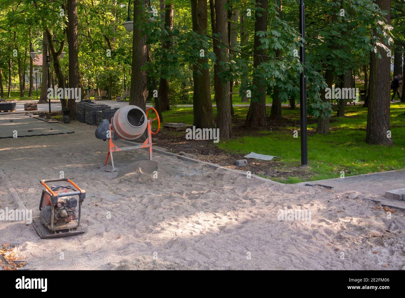 Die Reparatur des Gehwegs im Stadtpark. Betonmischer und Vibrationsplatte zum Verlegen von Pflasterplatten auf Sand. Selektiver Fokus. Stockfoto