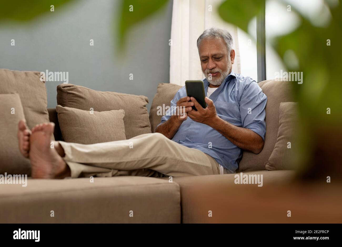 EIN ÄLTERER ERWACHSENER, DER BEQUEM AUF DEM SOFA SITZT UND BENUTZT MOBILTELEFON Stockfoto