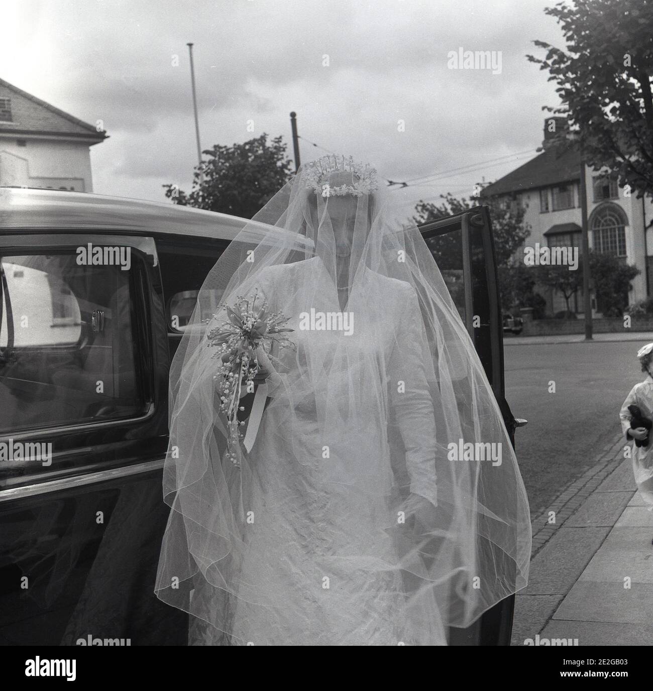 1960s, historisch, eine Braut in ihrem Hochzeitskleid und Kopfstück mit langem Schleier bedeckt ihr Gesicht, tritt aus ihrer Hochzeit oder Brautwagen, Ankunft für die cermony, England, Großbritannien. Der lang bedeckende Gesichtsschleier symbolisierte traditionell Bescheidenheit und Gehorsam und war eine elegante und allgemein getragene Brautkleider in dieser Zeit. Stockfoto