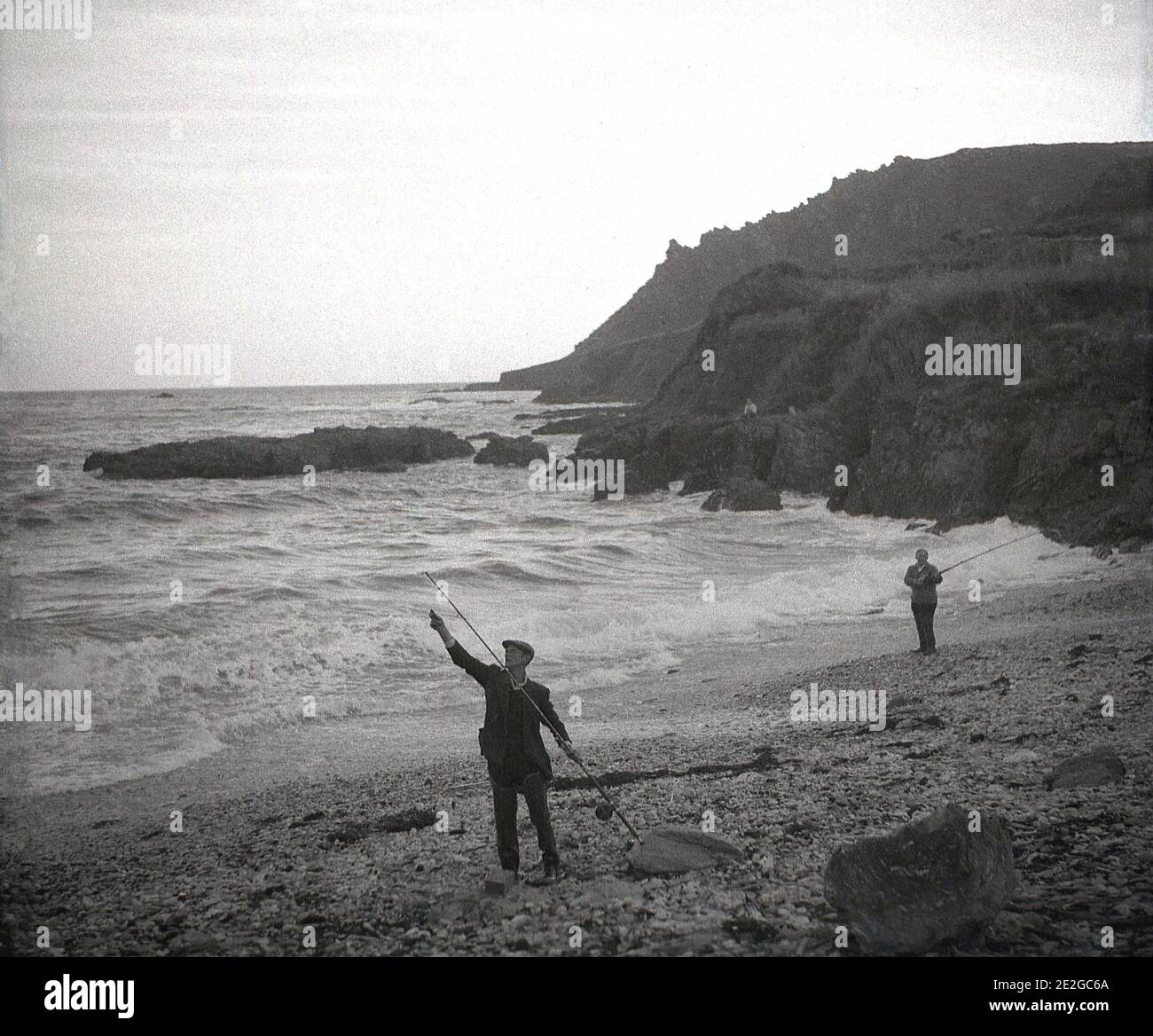 1960er Jahre, historisch, in einer felsigen Bucht am Meer, an einem steinigen Strand, zwei Männer in Watvögeln Seefischen, von einer Person in der Ferne sitzen auf den Klippen mit Blick auf die Bucht, England, Großbritannien beobachtet. Stockfoto