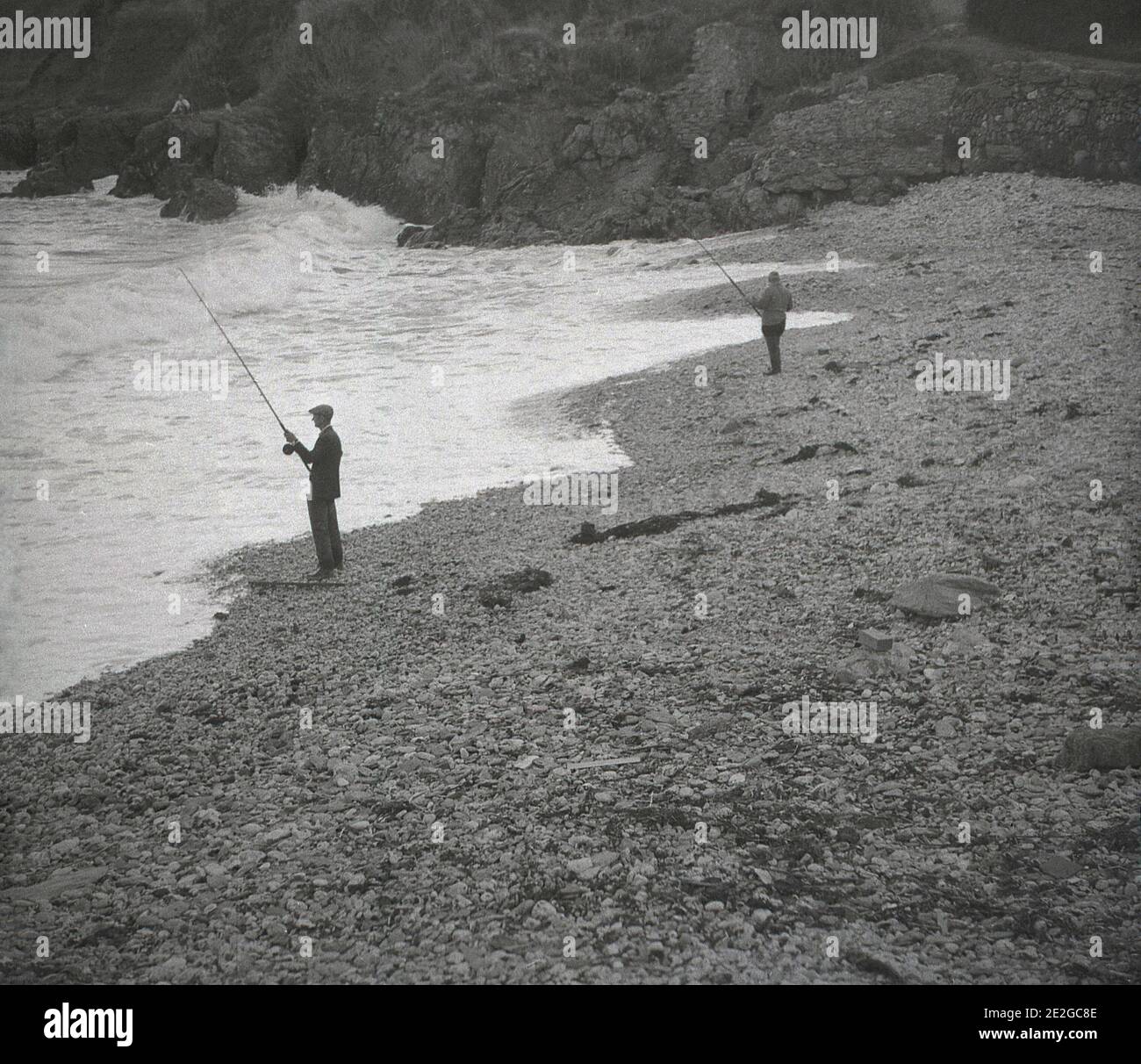 1960er Jahre, historisch, in einer geschützten Bucht, an einem steinigen Strand, zwei Männer in Watvögeln Seefischen, von einer Person in der Ferne sitzen auf den Klippen mit Blick auf die Bucht, England, Großbritannien beobachtet. Stockfoto