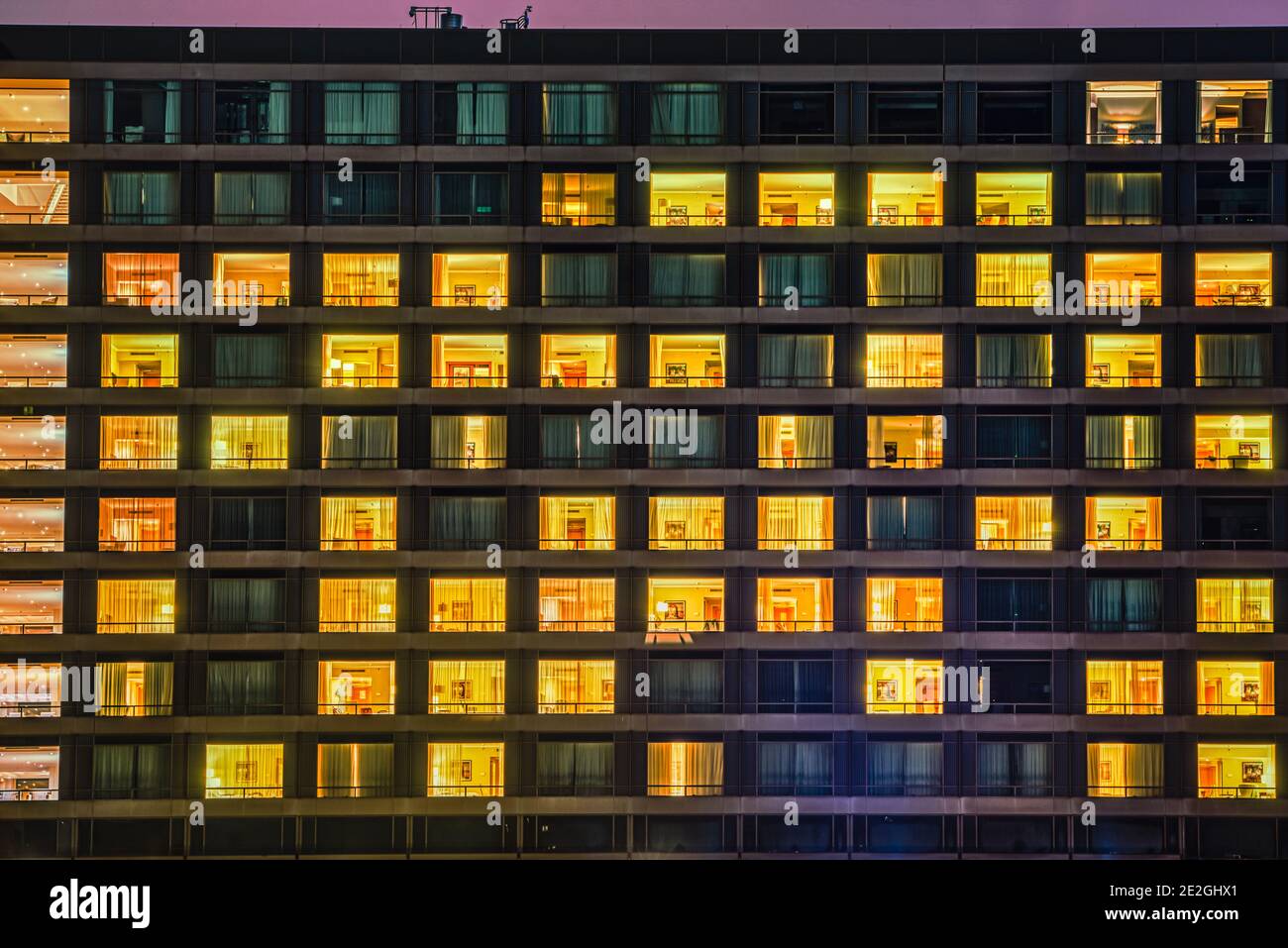Die Gebäudefassade zeigt nachts Fenster und Zimmermuster. Das Äußere zeigt beleuchtete und dunkle Wohnräume mit Menschen, die wie Nachbarn leben Stockfoto