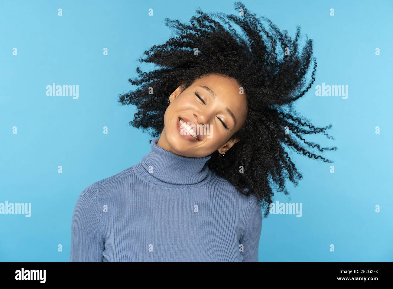 Glückliche Afro-Frau lächelt, hat gute Laune, tanzt mit ihrem schwarzen lockigen Haar fliegen, isoliert auf blau Stockfoto