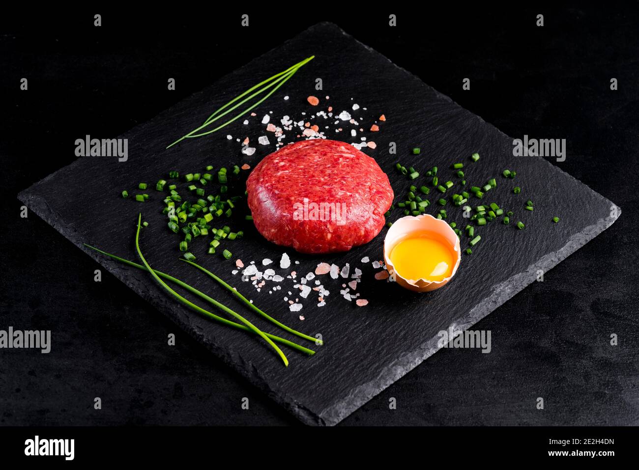 Rindertartar mit Ei auf einem schwarzen Steinteller serviert. Tartar auf schwarzem Teller mit Kräutern, Salz und Hühnerdotter. Rohes Tartarfleisch.Schwarzer Hintergrund. Oben Stockfoto