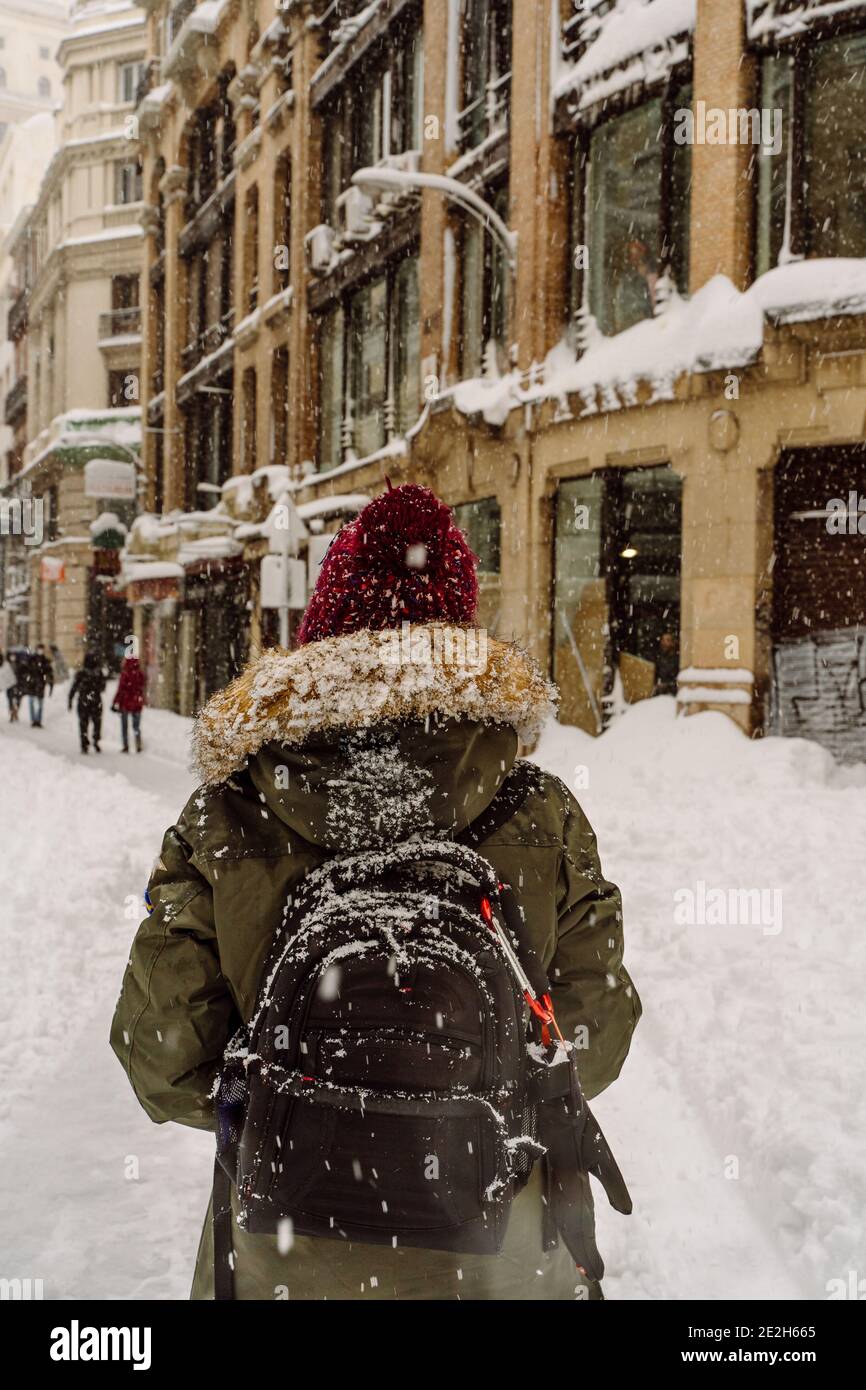 Frau in der verschneiten Stadt geht unter dem Schneesturm mit einem Rucksack, trägt einen Pelzhut und Mantel. Rückansicht Stockfoto