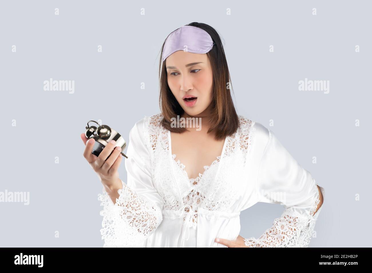 Asiatische Frau in einem weißen Satin Nachthemd und trägt Spitze Robe schockiert, als er aufwacht spät. Ein Mädchen, das spät aufwacht, schaut auf den Wecker auf einem Licht Stockfoto