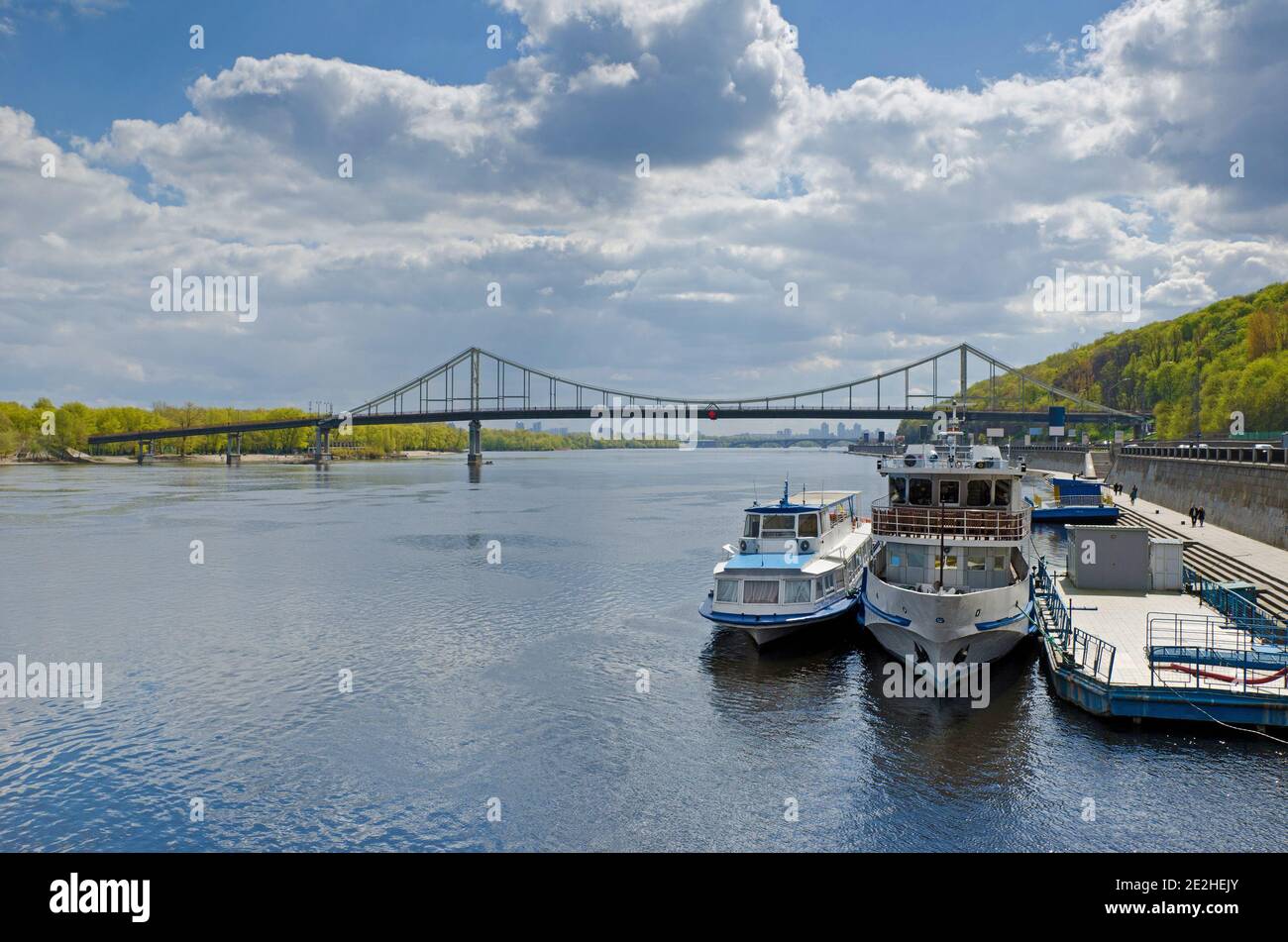 Fußgängerbrücke über den Fluss Dnipro und Böschung mit Touristenbooten in Kiew, Ukraine Stockfoto