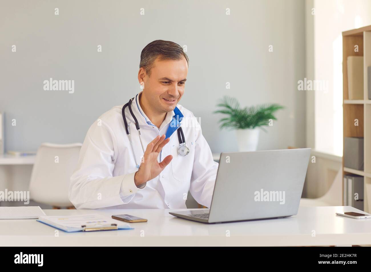 Lächelnder eHealth-Arzt winkt Hand an Laptop und grüßt Patient während eines Videoanrufs Stockfoto