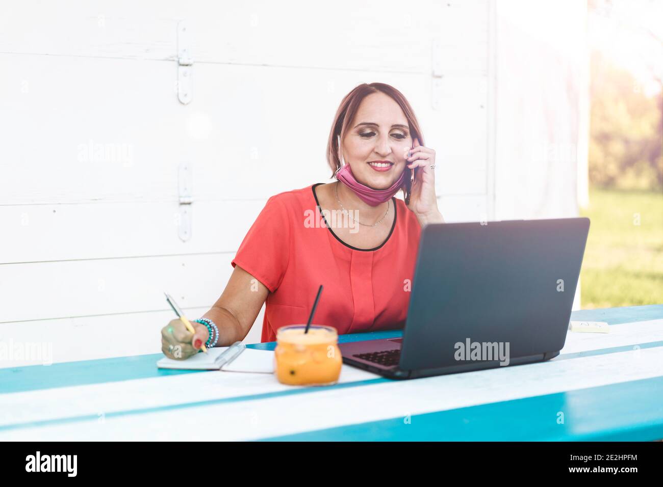 Lächelnde Frau 40 Jahre alt trägt offene Schutzmaske mit Laptop und Handy während des Endes des Coronavirus Ausbruch. Unternehmerin w Stockfoto