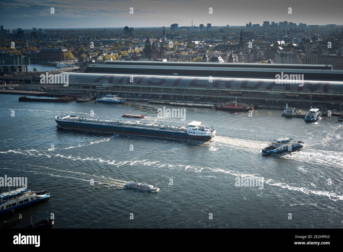 Die Stadt Amsterdam liegt über dem Hauptbahnhof und dem IJ-Kanal mit vorbeifahrenden Schiffen und Schiffen und Fähren. Stockfoto