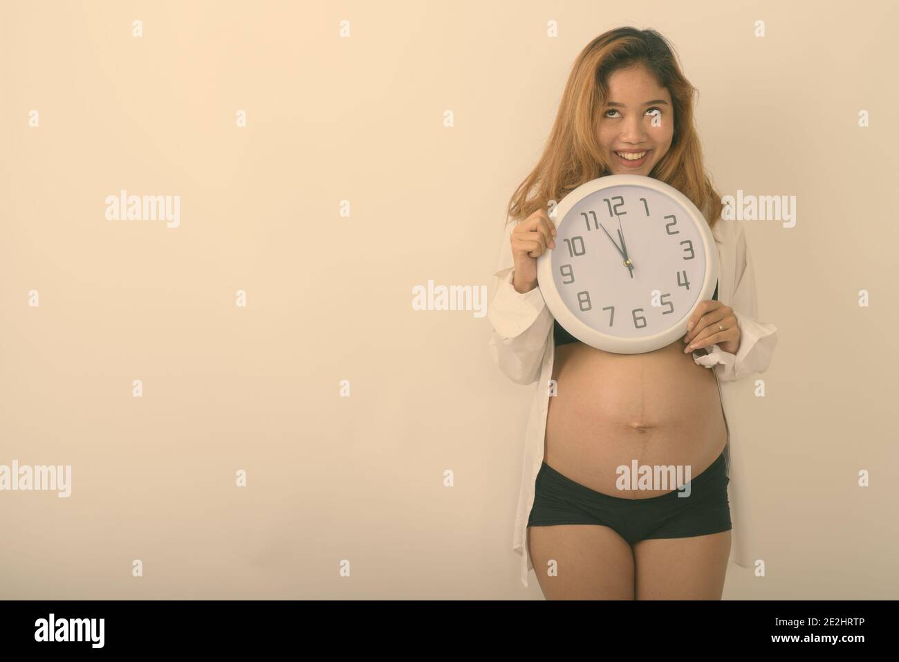 Studio shot von jungen asiatischen schwangere Frau glücklich lächelnd, während sie Wanduhr mit den Augen rollen vor weißem Hintergrund Stockfoto