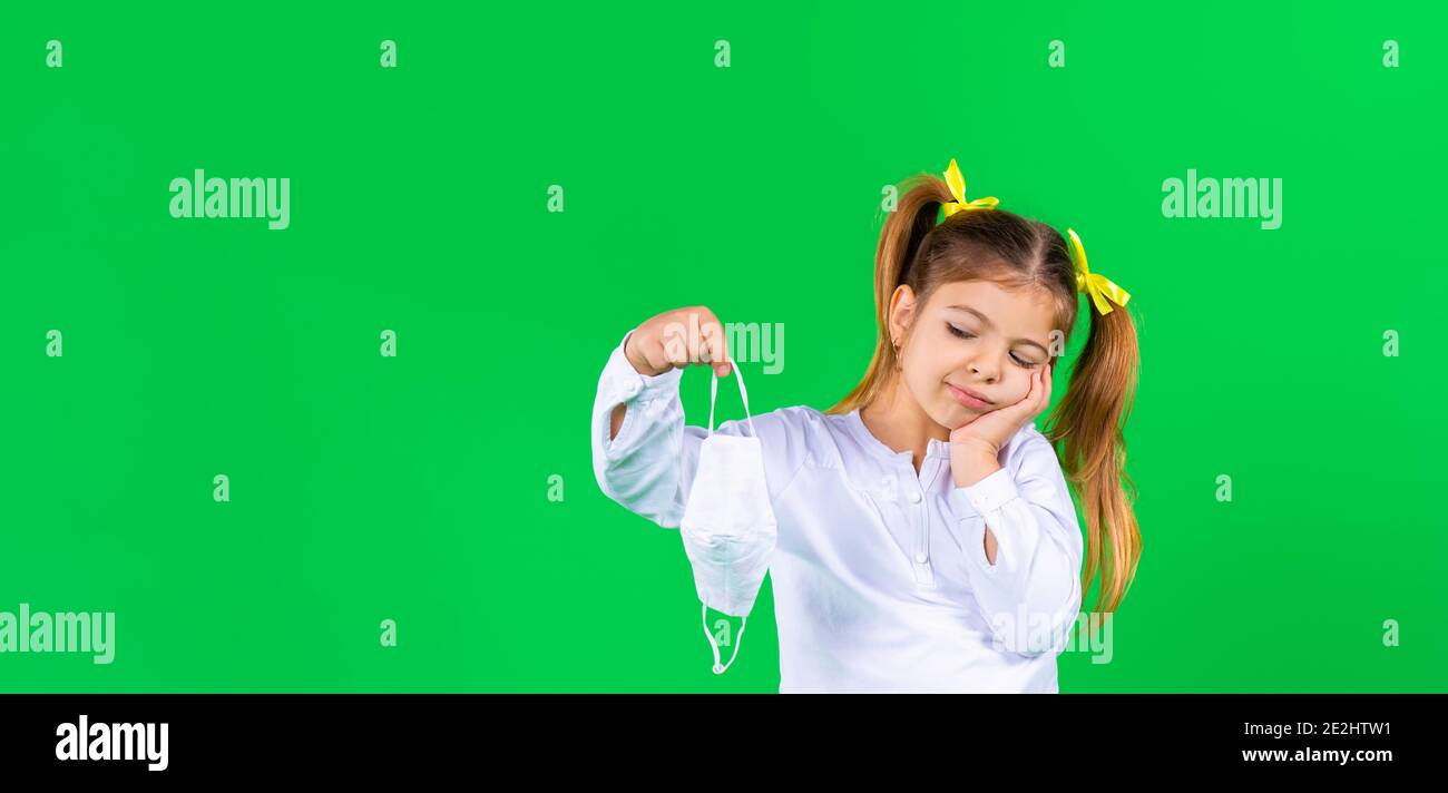 Banner. Ein nachdenkliches Mädchen im Vorschulalter hält eine medizinische Maske und sieht sie mit Unmut an. Grüner Hintergrund. Stockfoto