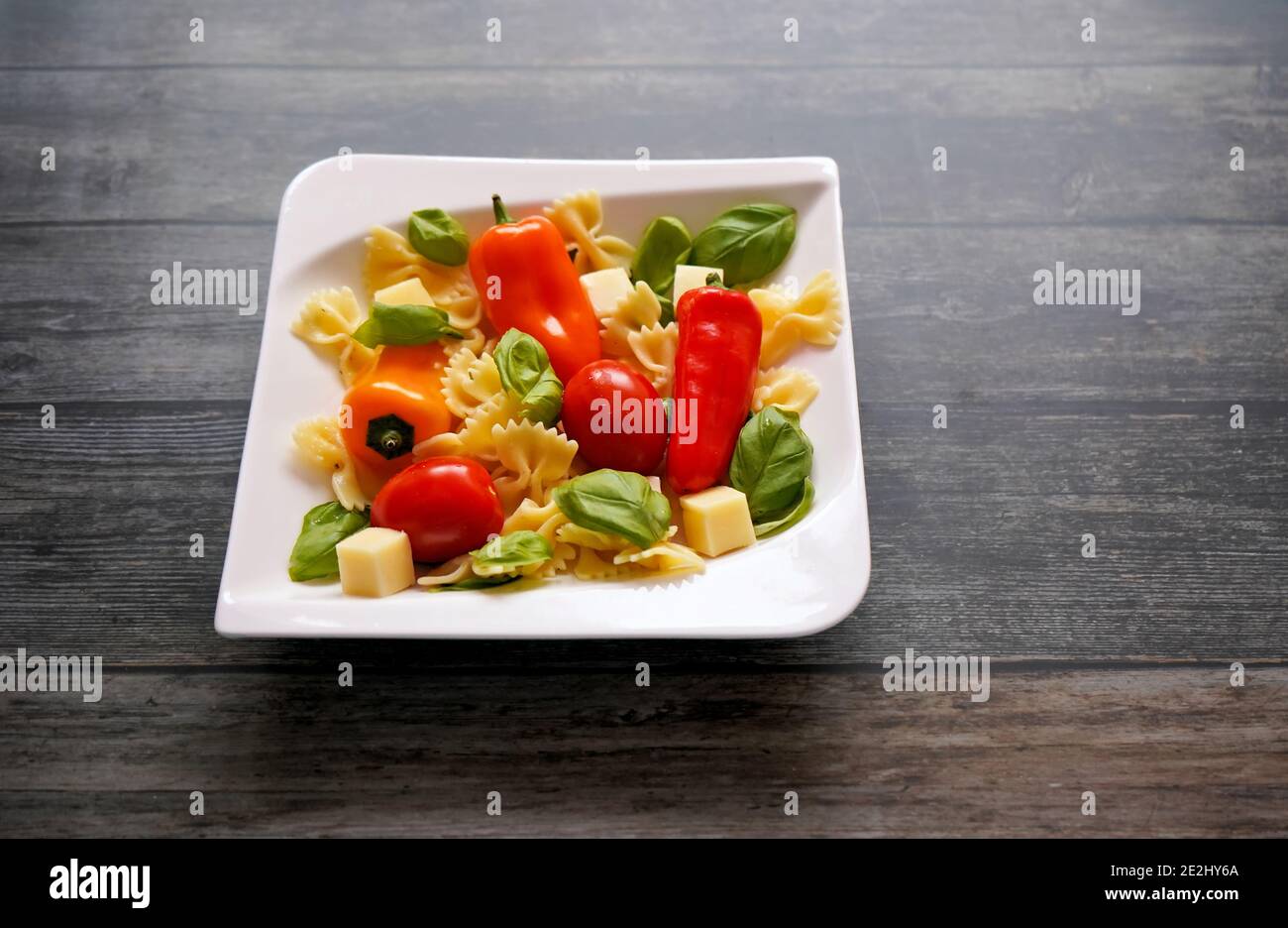 Frische Zutaten für einen gesunden Salat mit Nudeln, Tomaten, Paprika, Käsewürfeln und Basilikum auf einem weißen quadratischen Teller. Dunkler Holzhintergrund. Stockfoto