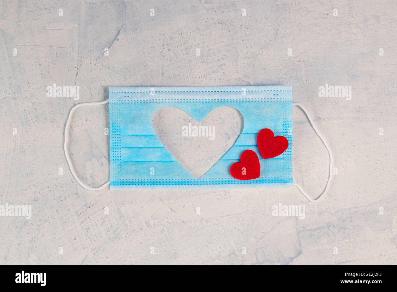 Medizinische Maske mit Ausschnitt Silhouette des Herzens und zwei roten Herzen auf weißem Stein Hintergrund. Valentinstag Hintergrund. Valentinstag-Konzept. Flach Stockfoto