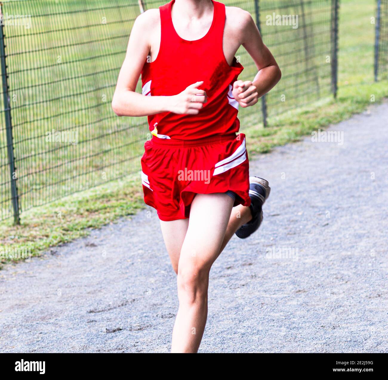 Vorderansicht eines sehr skinner High School Jungen, der während eines Cross Country Rennens auf Schotter läuft in roter Uniform mit weißen Streifen. Stockfoto