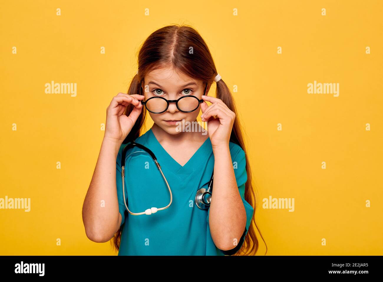 Mädchen trägt ein blaues medizinisches Outfit und ein Stethoskop, zukünftige Arzt, und zeigt auf ihrem Gesicht ernsthafte Emotion, Blick unter der Brille Stockfoto