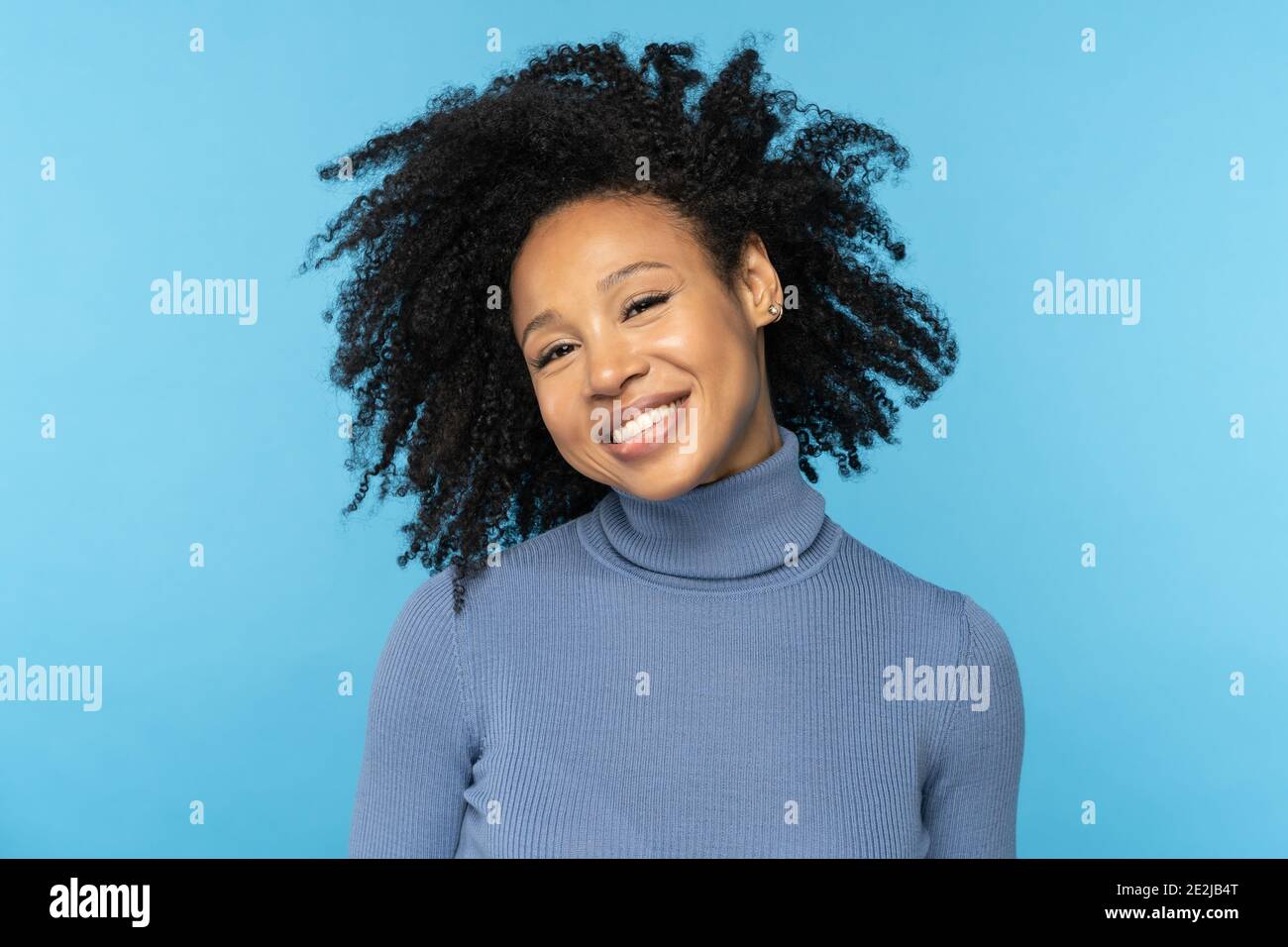 Glückliche Afro-Frau lächelt, hat gute Laune, tanzt mit ihrem schwarzen lockigen Haar fliegen, isoliert auf blau Stockfoto