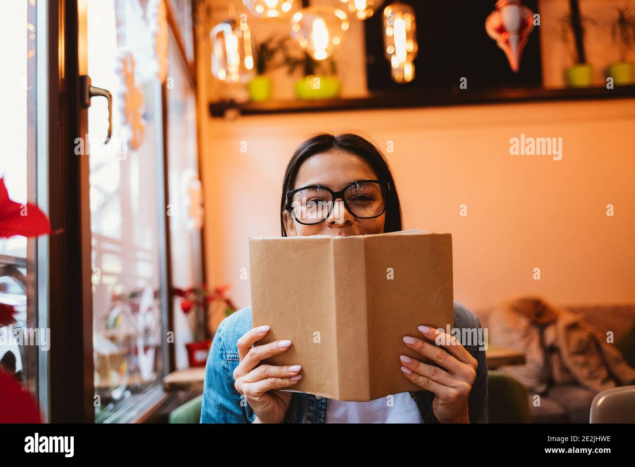Junge fröhliche Frau in Brille und Jeansjacke lesen ein Buchen Sie in einem Café und schauen Sie sich die Kamera an Stockfoto
