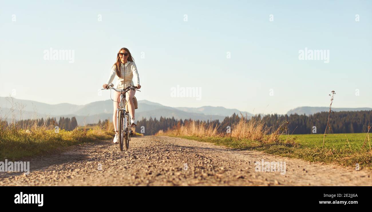 Junge Frau fährt Fahrrad auf staubigen Landstraße, lächelnd, verschwommen sonnigen Landschaft im Hintergrund. Breite Bannerfläche für Text rechts Stockfoto