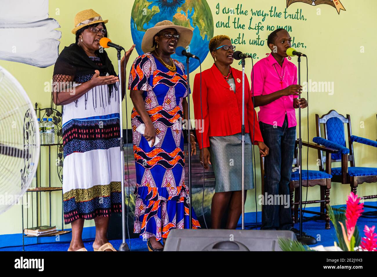 Drei Sängerinnen und ein Sänger stehen mit Mikrofonen vor einem gemalten Wandbild eines Globus und eines Bibelzitates auf der Bühne einer Baptistenkirche in The Lime, Grenada, und singen gemeinsam. Stockfoto