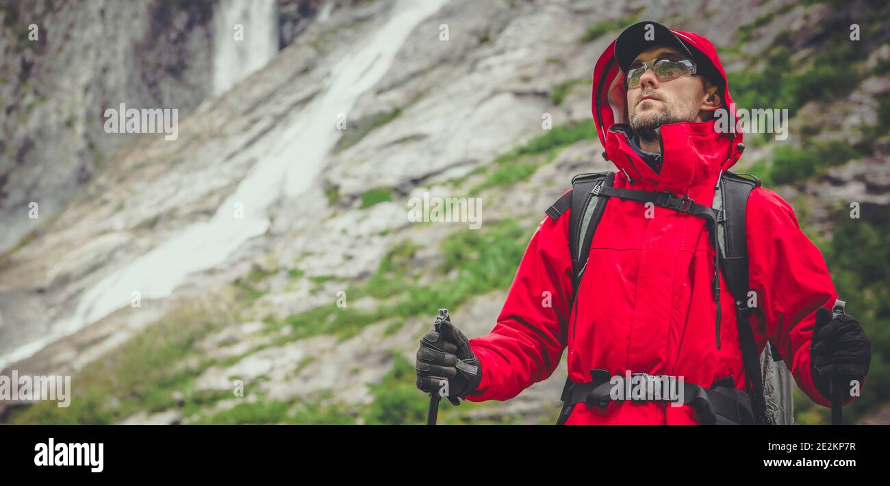 Outdoor Sport und Erholung Thema. Kaukasischer Wanderer in den 30er Jahren mit Red Raincoat und Safety Sport Brille auf dem Scenic Alpine Trailhead. Felsen und Stockfoto