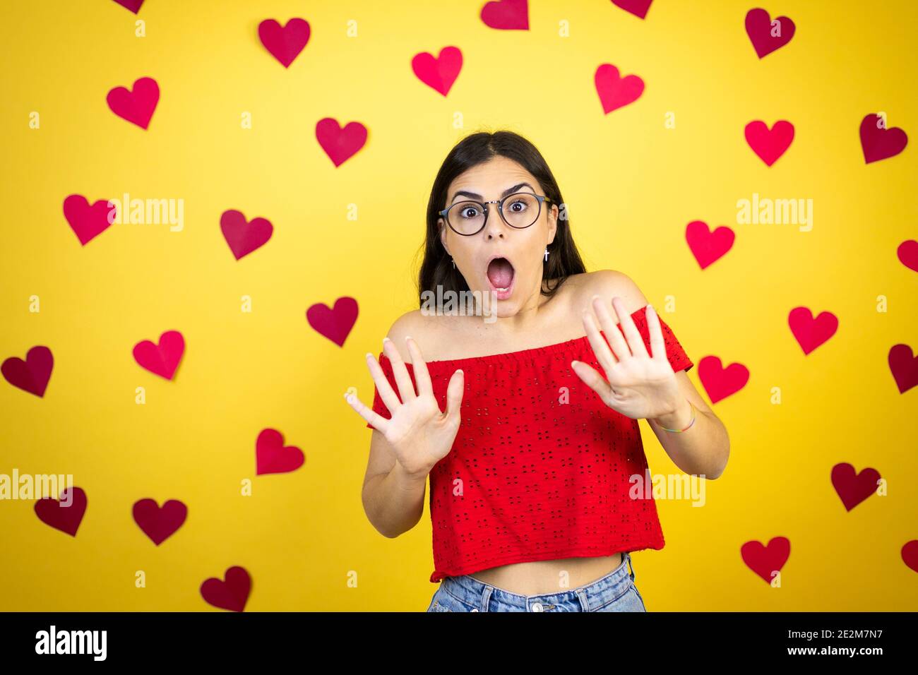 Junge schöne Frau auf gelbem Hintergrund mit roten Herzen Angst und erschrocken vor Angst Ausdruck Stop Geste mit Händen, schreien im Schock. Schwenken Stockfoto