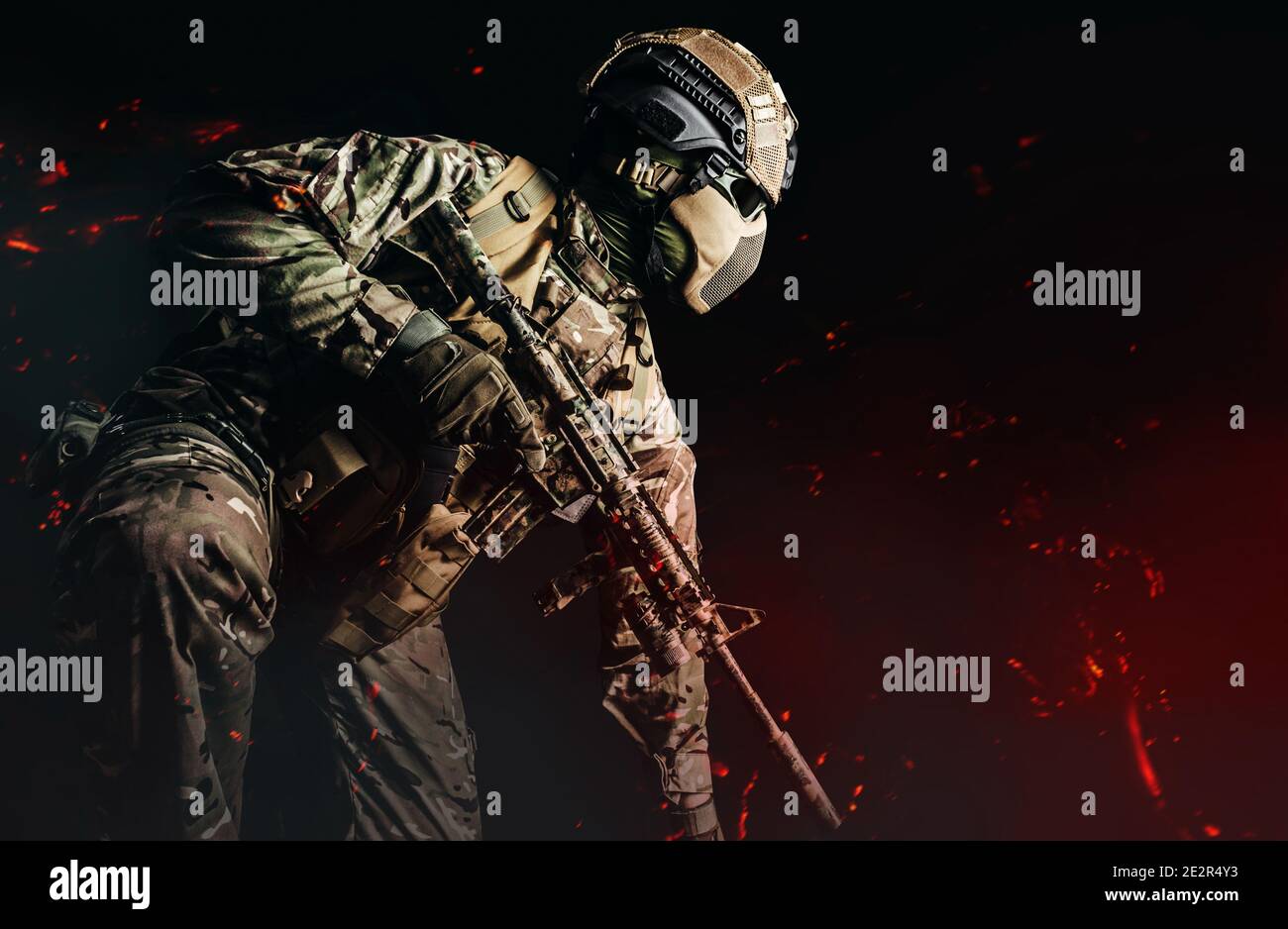 Foto eines voll ausgerüsteten Soldaten in gepanzerter Weste, Helm, Gesichtsbrille und Schutz kniend und Angriff mit Gewehr auf dunklem Hintergrund mit Asche Stockfoto