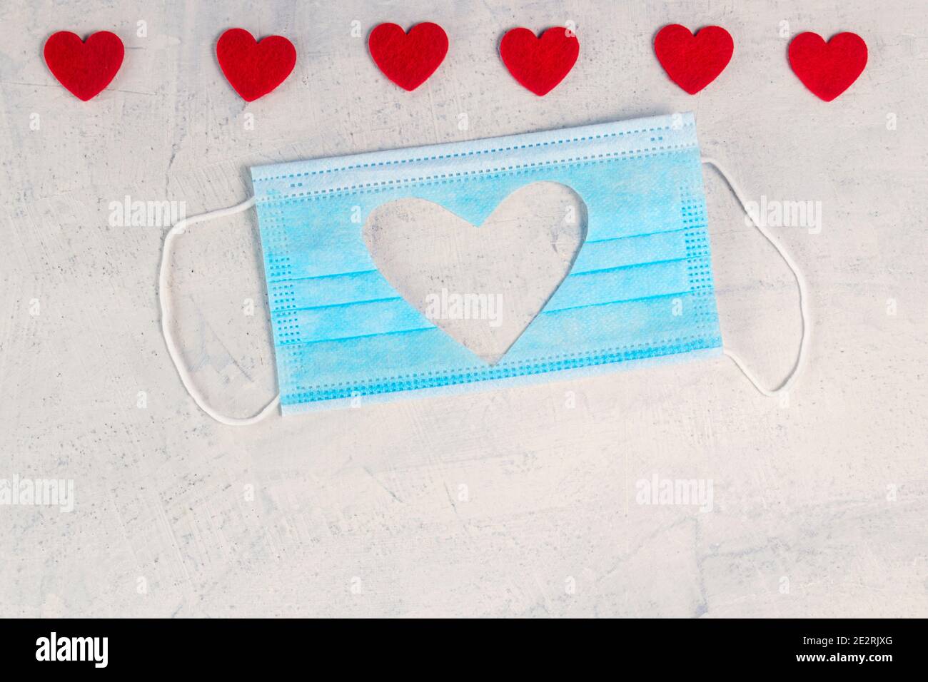 Medizinische Maske mit Ausschnitt Silhouette von Herzen und roten Herzen auf weißem Stein Hintergrund. Valentinstag Hintergrund. Valentinstag-Konzept. Flach liegend. Stockfoto