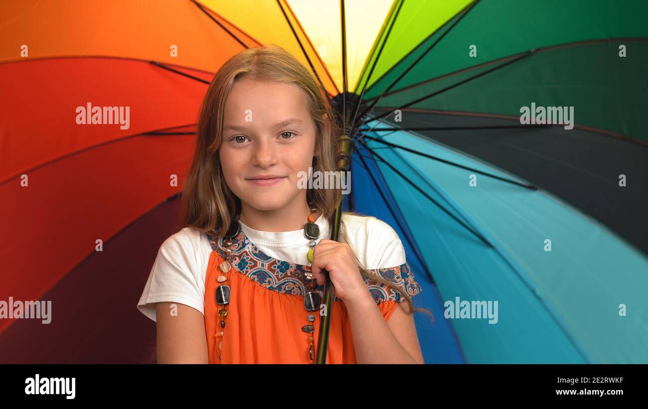 Charmante kleine Mädchen in orange Kleid hält Regenbogenfarben Regenschirm Stehen und jubeln im Studio, während man auf die Kamera schaut Stockfoto