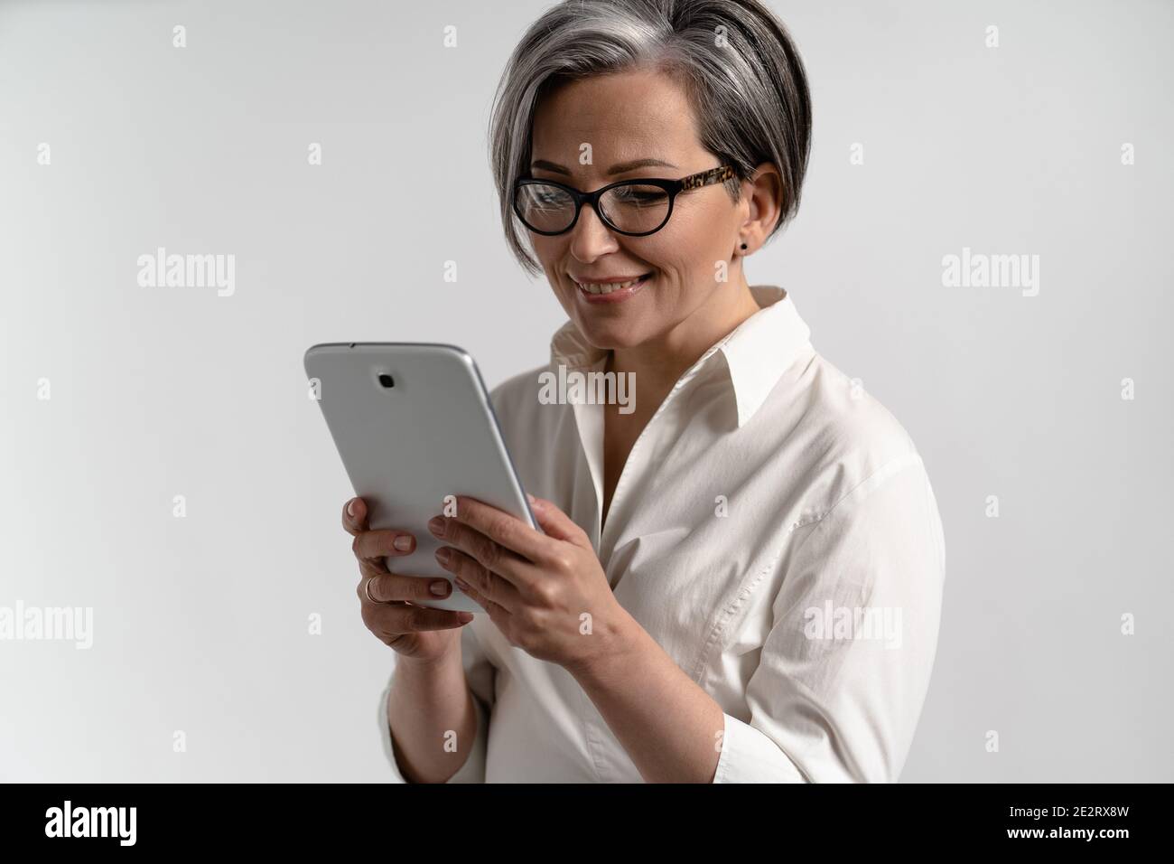 Porträt einer grauhaarigen älteren Frau in weißem Hemd und Brille mit digitalem Tablet. Technologie durch ältere Menschen, die im Internet einkaufen Stockfoto